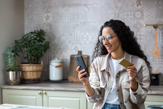 A woman in her kitchen paying her bills online using her debit card and her smartphone