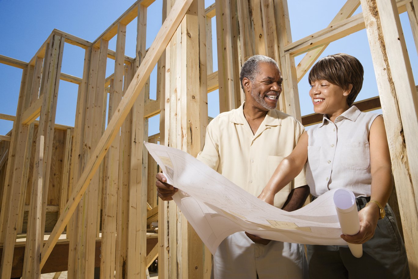 Married couple happily reviewing blueprints in front of their new-construction home being framed