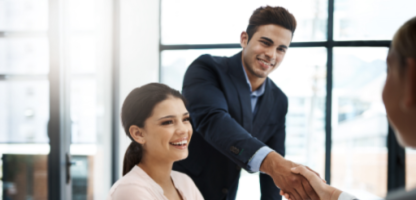 Two people sitting at a table in a bright office, with one person standing and shaking hands with someone across the table