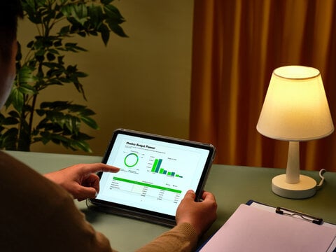 Person reviews their budget on a tablet, while sitting at a desk with a clipboard and small lamp